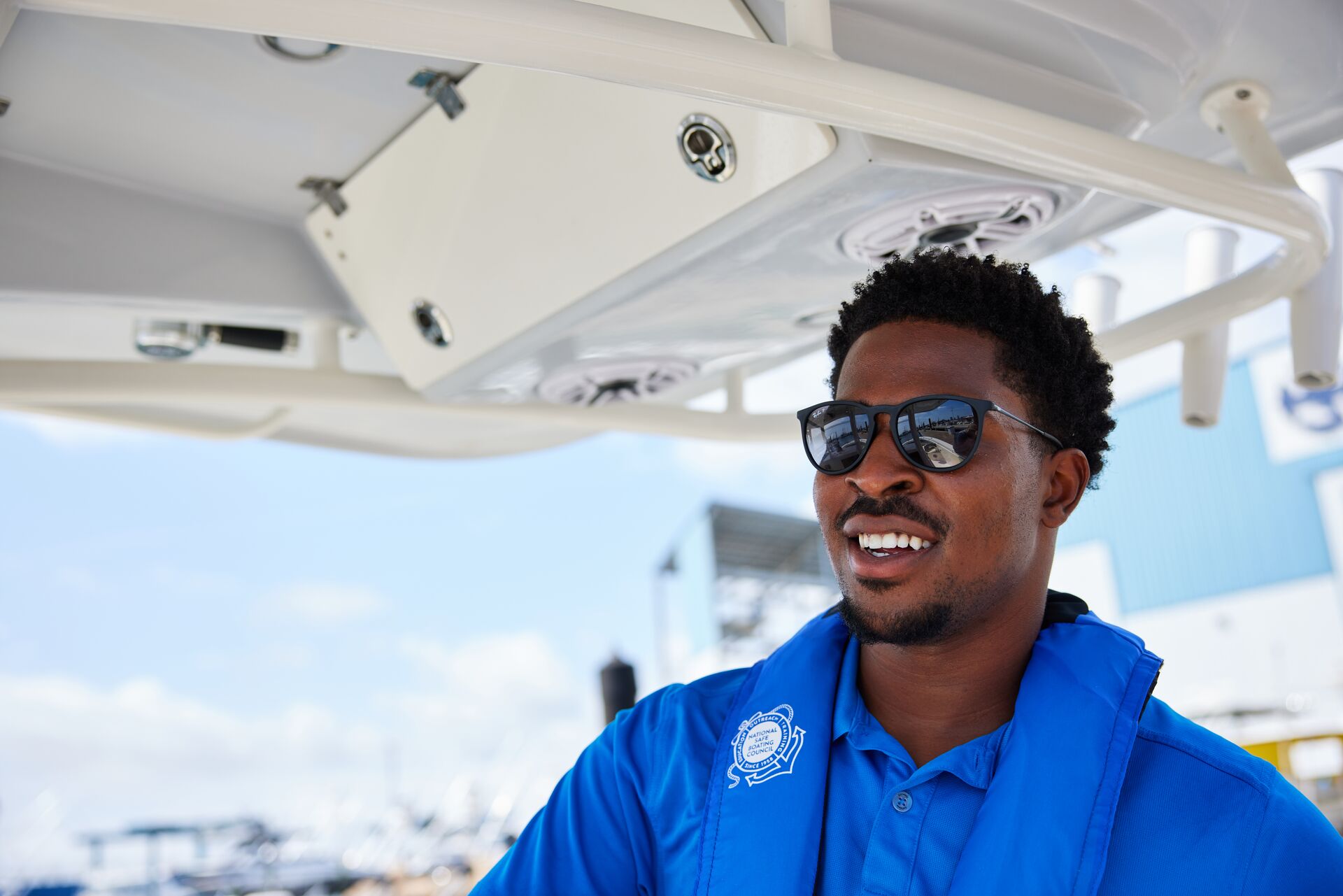 A man in a blue shirt smiles, wears a life jacket, drives a boat. 