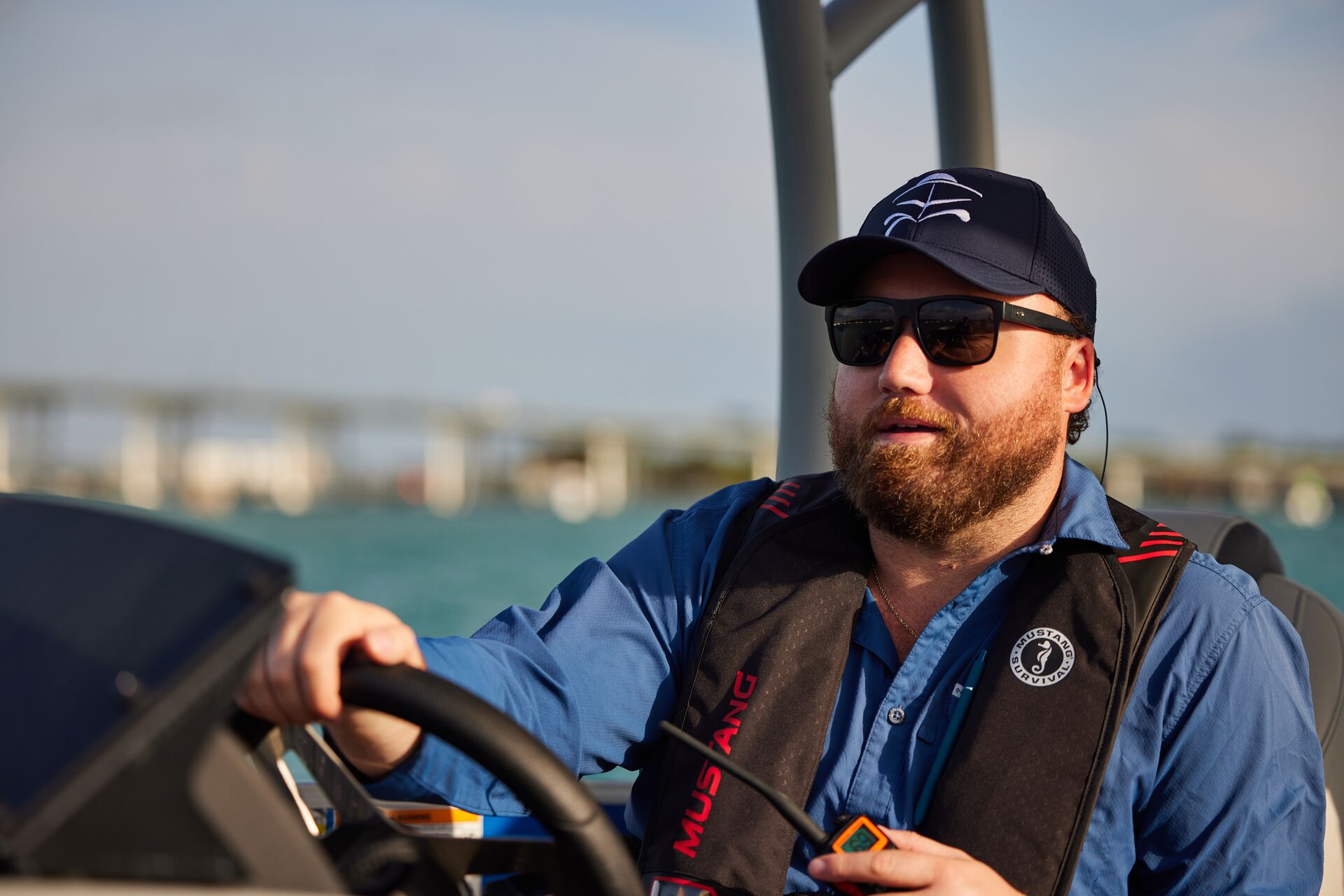 A man drives a boat while holding a radio and wearing an inflatable life jacket.
